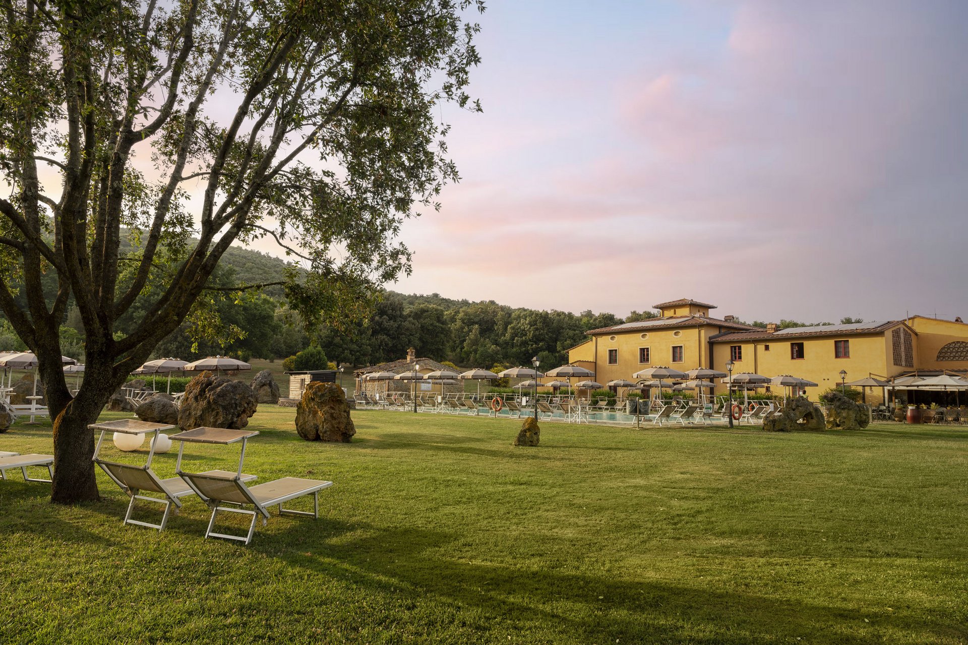 Roadbike Holidays © Hotel Casolare Le Terre Rosse Grüner Garten mit Bäumen, Liegestühlen und Sonnenschirmen neben einem gelben Gebäude