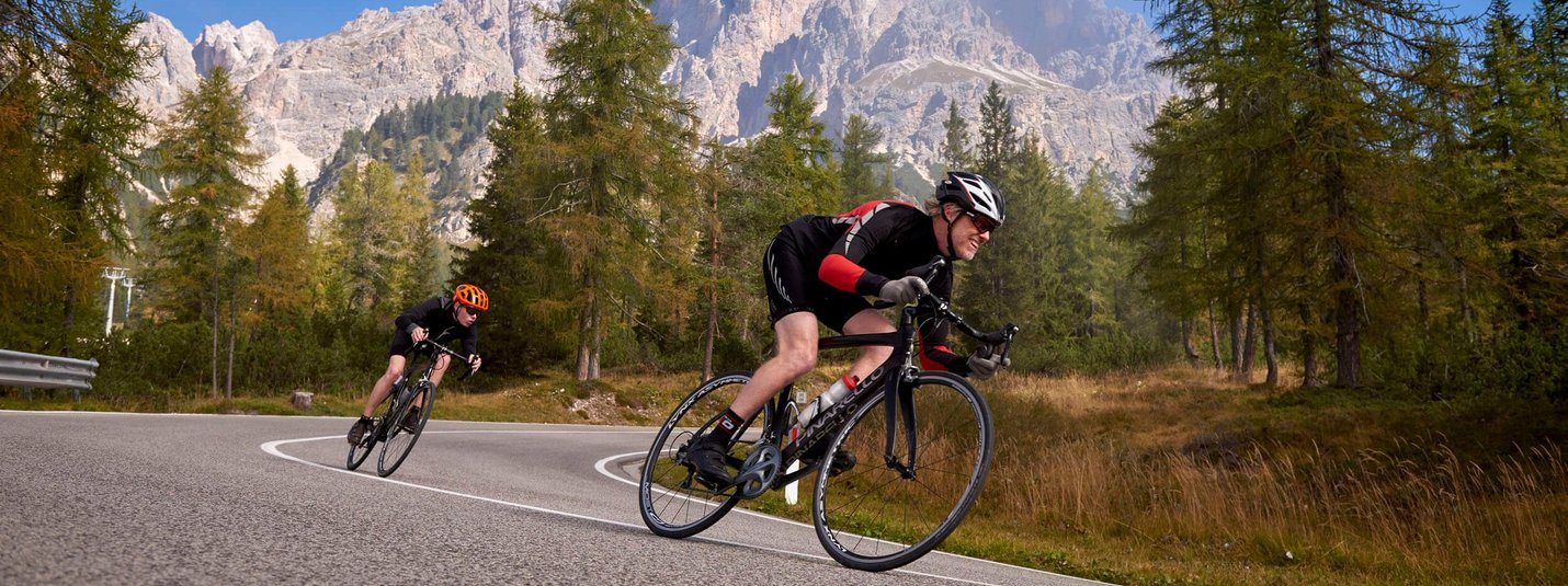 Rennrad Urlaub in der Region Drei Zinnen - Dolomiten © Joris Lugtigheid Zwei Radfahrer fahren auf kurviger Bergstraße mit Bäumen und Bergen im Hintergrund