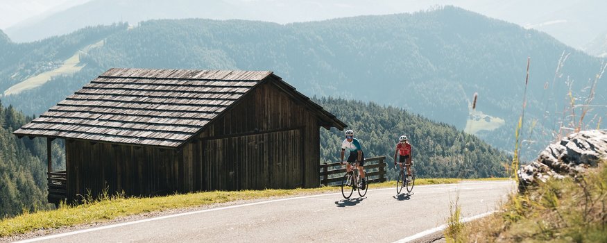 Sporthotel Exclusive **** © Alex Moling Two cyclists riding on mountain road next to wooden hut in the Alps