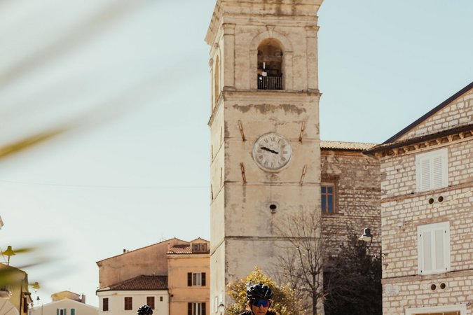 Roadbike Holidays © Alex Moling Zwei Radfahrer fahren durch eine sonnige Stadt mit einem alten Uhrturm