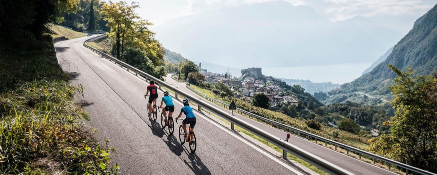 Noordelijke Gardameer © Alex Moling Drie wielrenners rijden op bergweg met uitzicht op dorp en meer