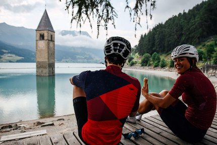 3 paesi Giro © Felix Saller Due ciclisti seduti vicino a un lago con una torre della chiesa sommersa