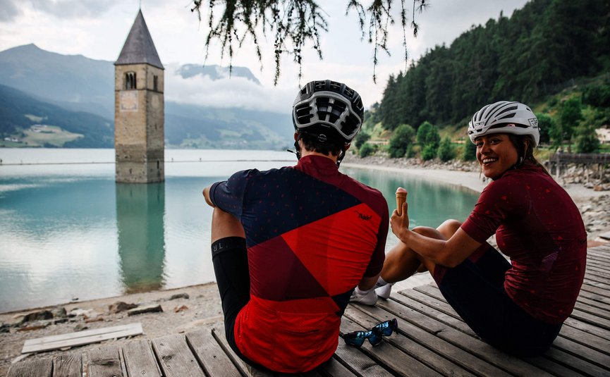 Vacanze in bici da corsa nella regione di Nauders – Lago di Resia © Felix Saller Due ciclisti seduti vicino a un lago con una torre della chiesa sommersa