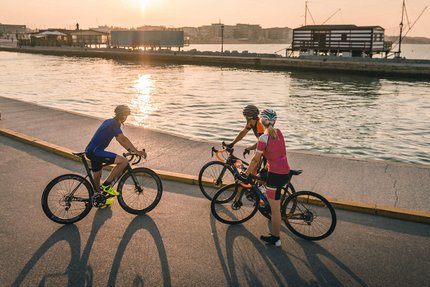 Nove Colli 130 © Alex Moling Three cyclists stopped by water at sunset with buildings in background
