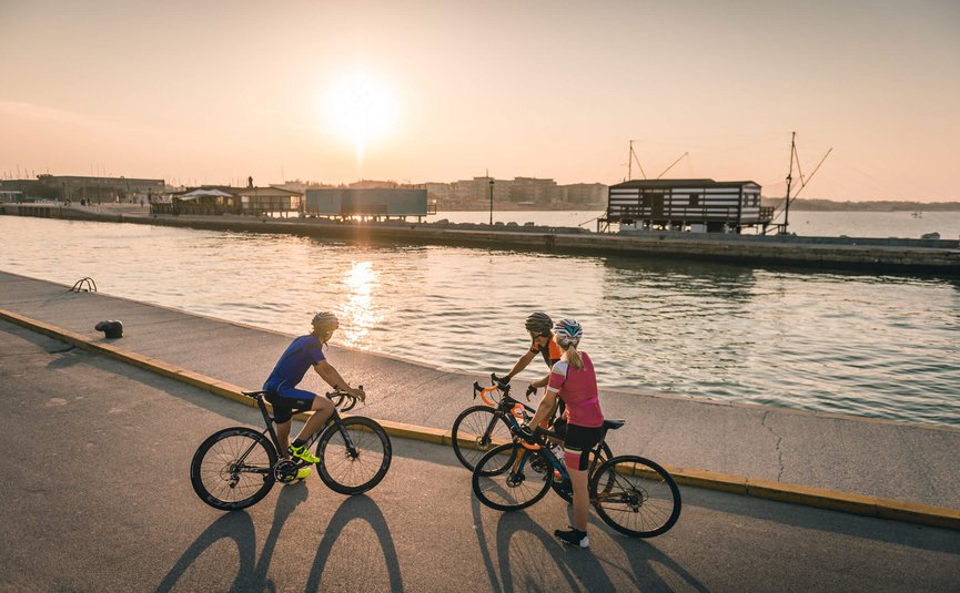 Road bike holidays on the Adriatic coast © Alex Moling Three cyclists stopped by water at sunset with buildings in background