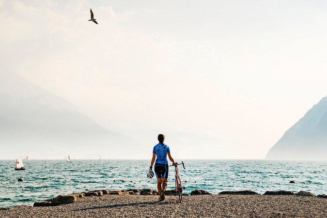 Roadbike Holidays © Alex Moling Fahrradfahrer am Seeufer mit Bergen und Segelbooten im Hintergrund