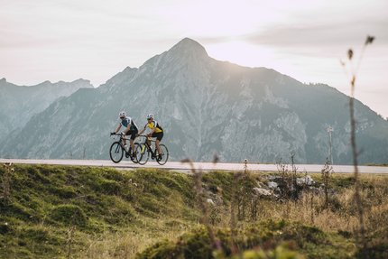 Postalm - Rundweg über Hallein © SalzburgerLand - Stefan Schopf for WOM Medien Zwei Radfahrer fahren auf Bergstraße mit großem Berg im Hintergrund bei Sonnenuntergang