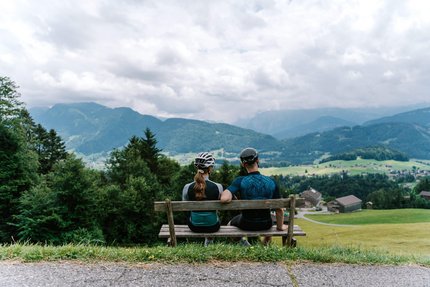 Ein Rennrad-Wochenende im Bregenzerwald © Bregenzerwald Tourismus - Christina Neubauer Zwei Radfahrer auf Bank blicken auf Berglandschaft mit Häusern und Wiesen