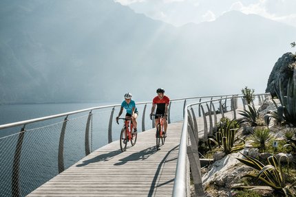 Trentino © Alex Moling Zwei Radfahrer auf einem Steg am See mit Bergen im Hintergrund
