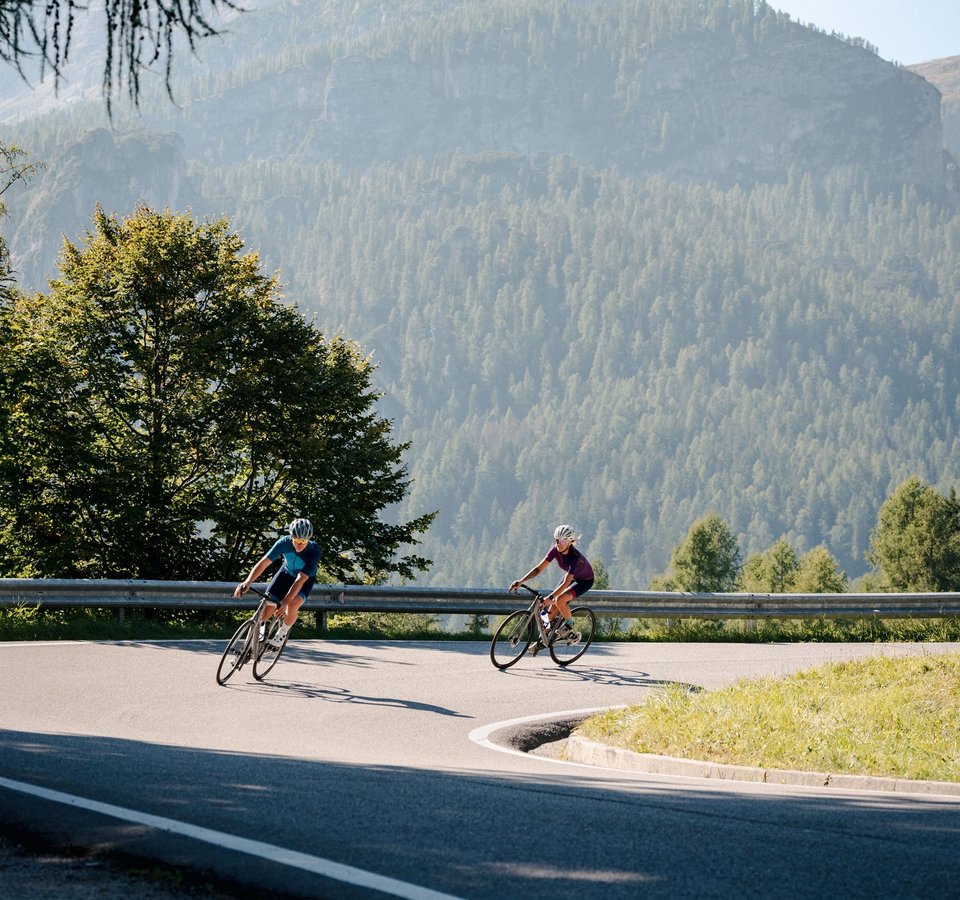 Italiens Pässeparadies © Josef Plaickner Zwei Radfahrer fahren auf kurviger Bergstraße mit bewaldeten Bergen im Hintergrund