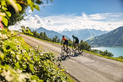 SalzburgerLand © SalzburgerLand Tourismus - WOM Medien Zwei Radfahrer fahren auf Bergstraße mit Blick auf See und Berge bei sonnigem Wetter