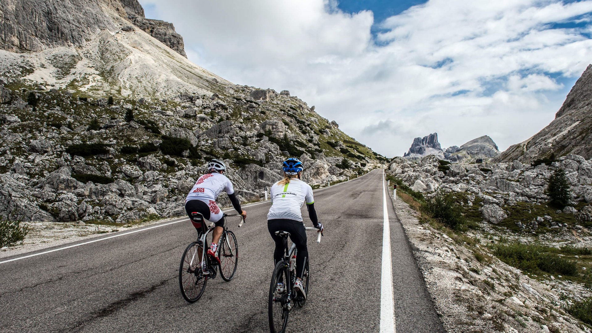 Roadbike Holidays © Paola Finali Zwei Radfahrer fahren eine Bergstraße mit Felsen und wolkigem Himmel.