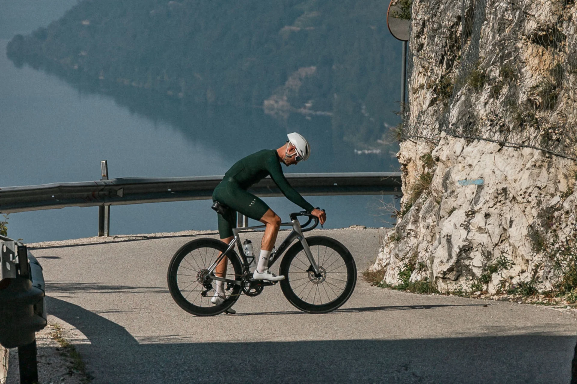 Roadbike Holidays © marc.moves x velysiped Radfahrer fährt auf bergiger Straße nahe Tunnel mit Seeblick