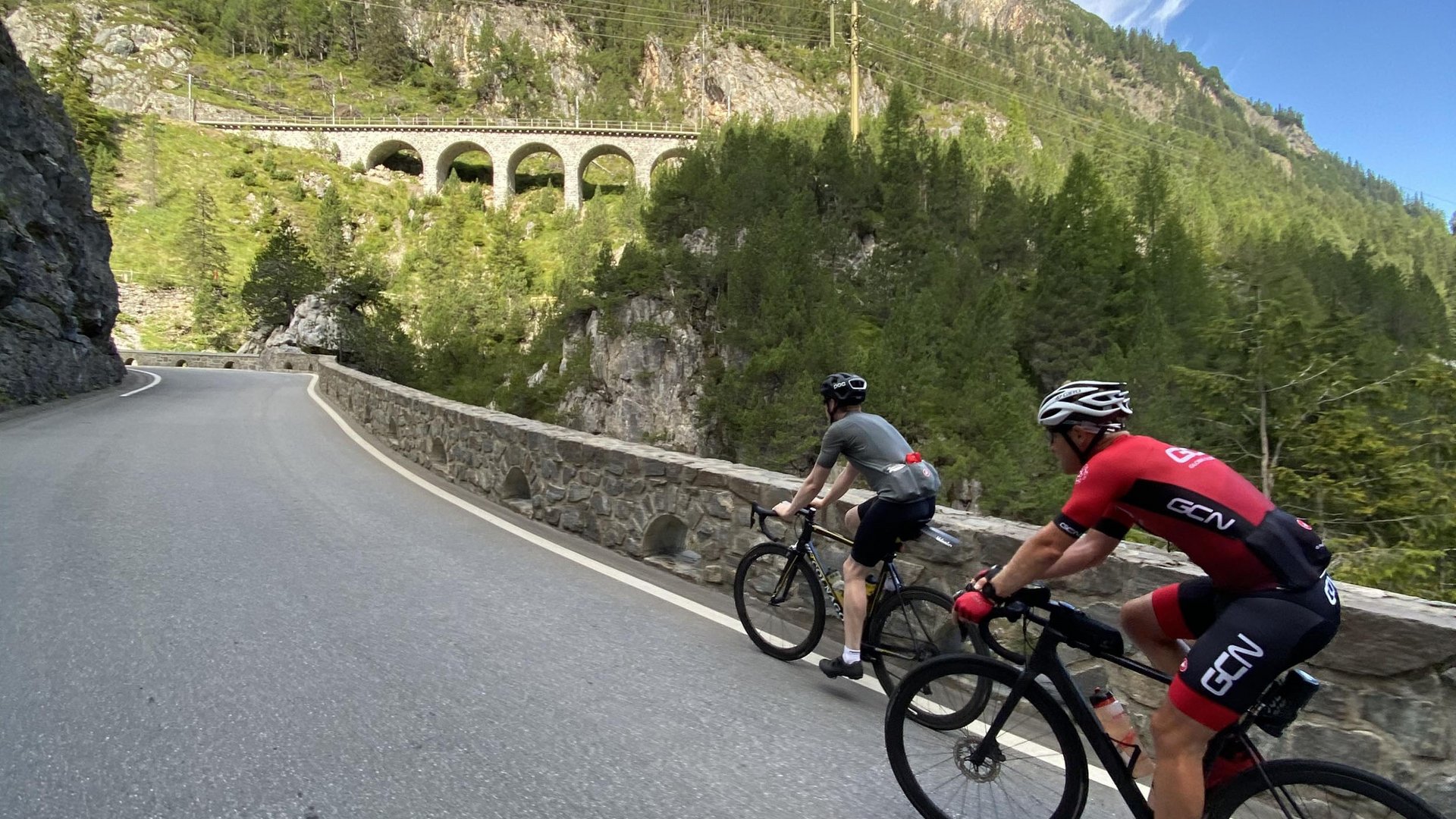 Roadbike Holidays © quäldich.de GmbH Zwei Rennradfahrer fahren bergauf auf einer kurvigen Straße mit Brücke und Wald