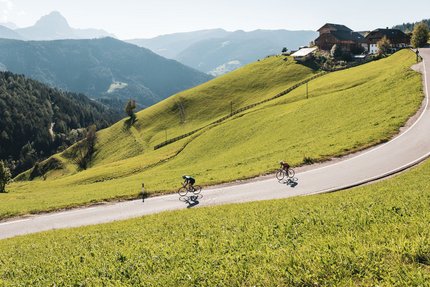 Funes and Val Gardena © Alex Moling Two cyclists ride a winding mountain road surrounded by green hills