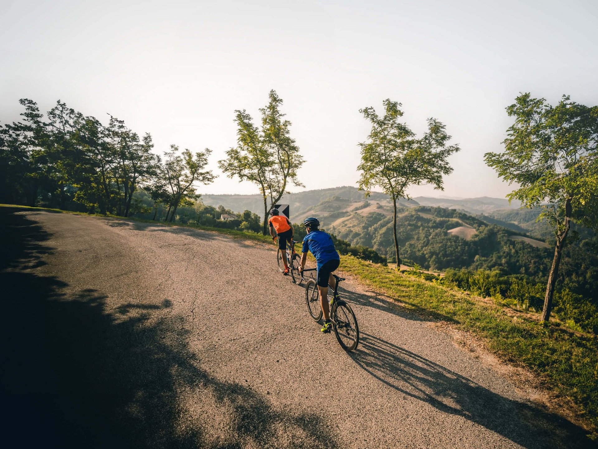 Roadbike Holidays © Alex Moling Zwei Radfahrer fahren eine kurvige Straße entlang mit Bäumen und Hügeln im Hintergrund