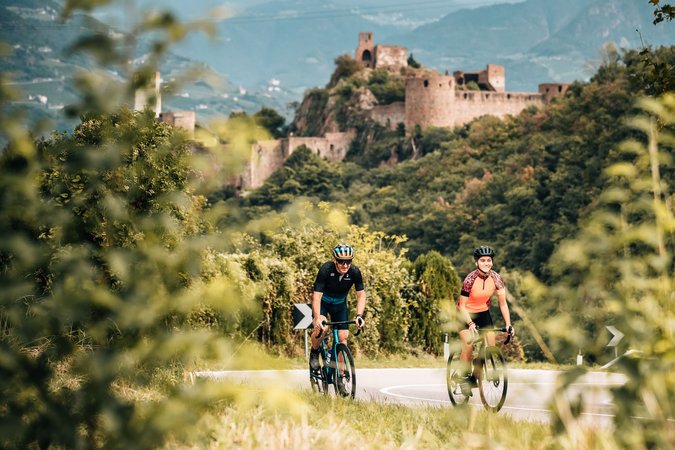 Roadbike Holidays © Jens Scheibe Zwei Radfahrer auf Straße vor einer Burg in bewaldeter Landschaft