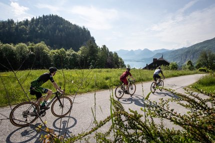Hausrunde - Rundweg über Mondsee © Erwin Haiden Drei Radfahrer auf einer Landstraße in einer bergigen Landschaft bei Sonnenschein
