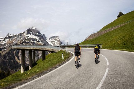 Vorarlberg © Bregenzerwald Tourismus - Merlijn Spenkerlink Zwei Radfahrer fahren eine Bergstraße mit schneebedeckten Gipfeln im Hintergrund