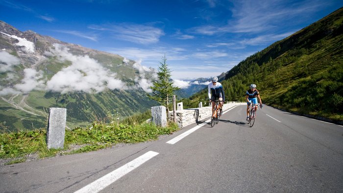 Tours & Travels © Heiko Mandl Two cyclists riding on a mountain road with mountains and blue sky in the background