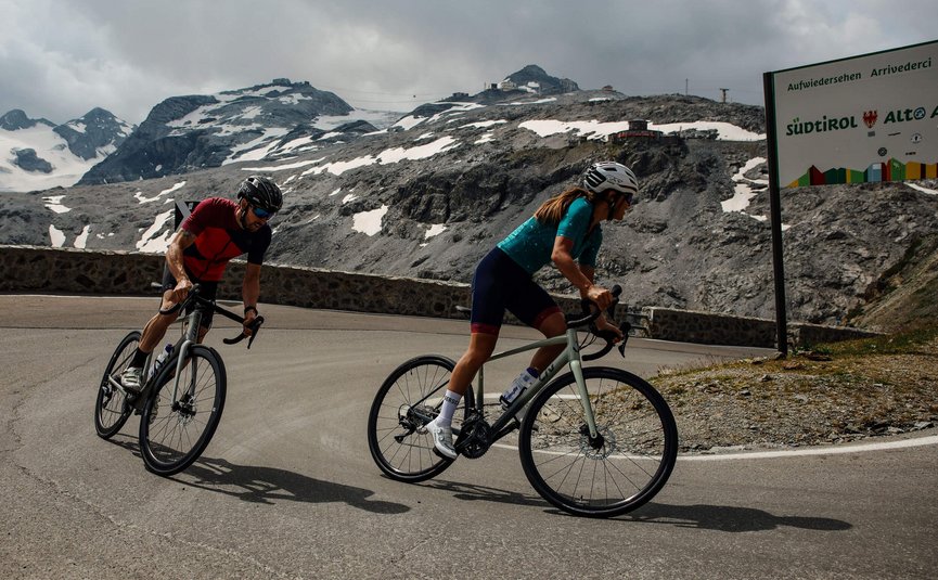 Vacanze in bici da corsa nella regione di Nauders – Lago di Resia © Felix Saller Due ciclisti su strada di montagna con vette innevate e cartello Alto Adige