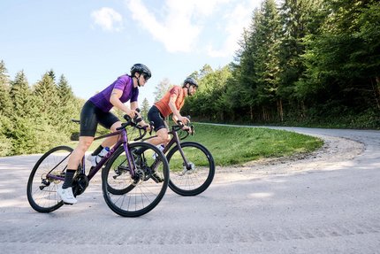 Bayern © Tobias Köhler Zwei Radfahrer auf der Straße in einer bergigen Waldlandschaft.